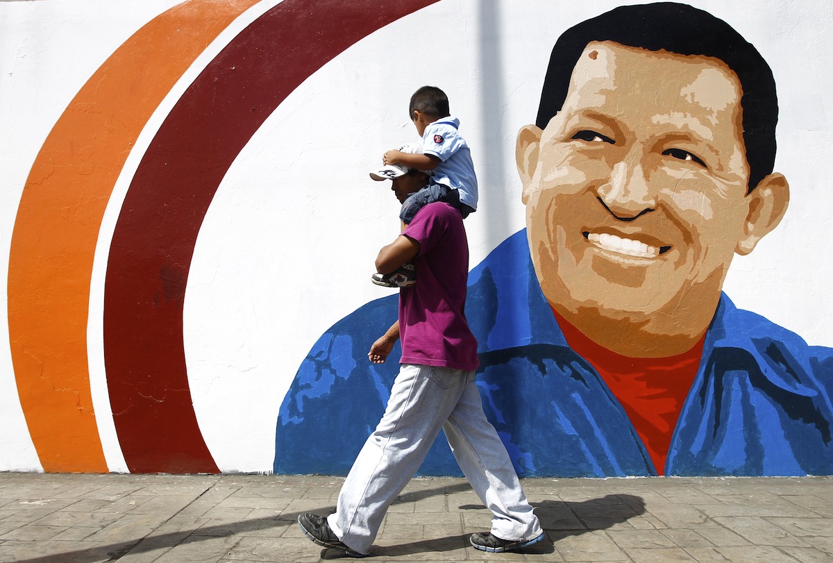 A man carrying a child walks past a mural of Venezuela's President Hugo Chavez in Caracas June 29, 2012. Chavez has a strong lead in most opinion polls as he seeks to beat opposition challenger Henrique Capriles and win a new six-year term in an October 7 election. REUTERS/Carlos Garcia Rawlins (VENEZUELA - Tags: POLITICS ELECTIONS)