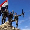 Militares sirios en la cima de una roca con armas y la bandera de Siria.