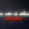 Letrero de neon en una pares oscura. En luz neo blanca se lee en letra cursiva "Cien años de soledad" y abajo en luz neon roja, entre corchetes en mayúscula se lee "NO REALMENTE".