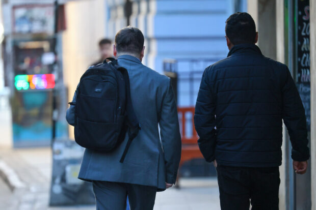 Dos hombres se ven caminando de espaldas. Uno viste traje gris y tiene una mochila en su hombro. El otro viste chaqueta negra.