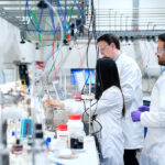 Universidad, ciencias sociales y democracia: una defensa común Una científica opera maquinaria con cables de colores mientras dos colegas supervisan el proceso frente a estantes con frascos químicos.