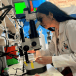 Investigar es trabajar Una científica con bata blanca de la Universidad de Chile analiza muestras bajo un microscopio profesional en un laboratorio equipado con instrumentos de alta precisión.