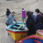 Un grupo de mujeres recolectoras y pescadoras artesanales trabaja junto a embarcaciones de madera pintadas de amarillo y rojo en una playa.